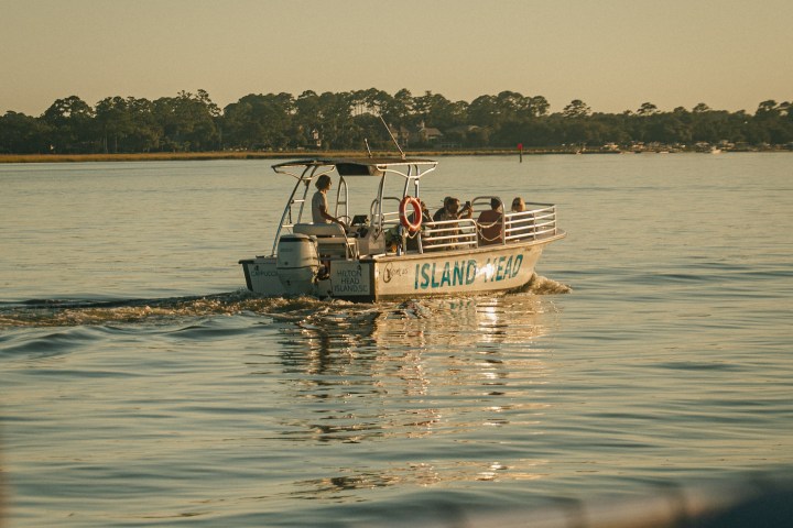 Boat with people on calm water during sunset, distant trees in background.