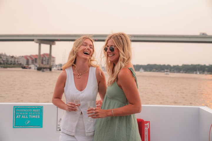 Two women smiling on a boat with a bridge in the background.