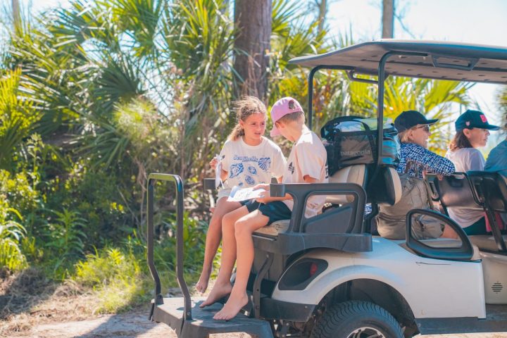Children sit on a parked golf cart, surrounded by lush greenery.