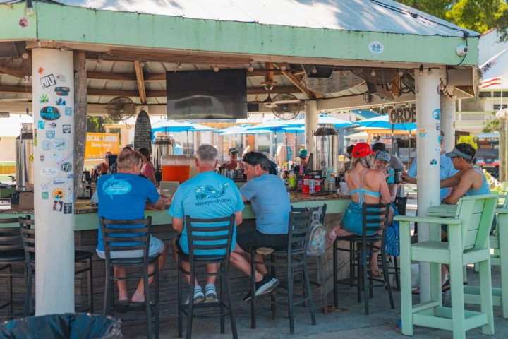 People sitting at an outdoor beach bar with a tiki-style roof and tall stools.