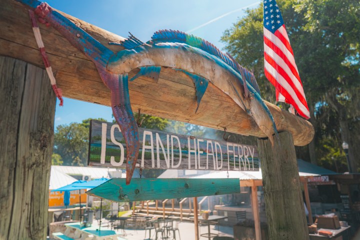 Wooden ferry sign with colorful fish sculpture, surrounded by trees and a U.S. flag under a clear blue sky.