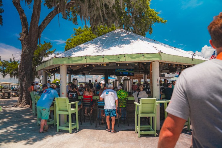 Outdoor bar with people seated and standing under a white pavilion on a sunny day.