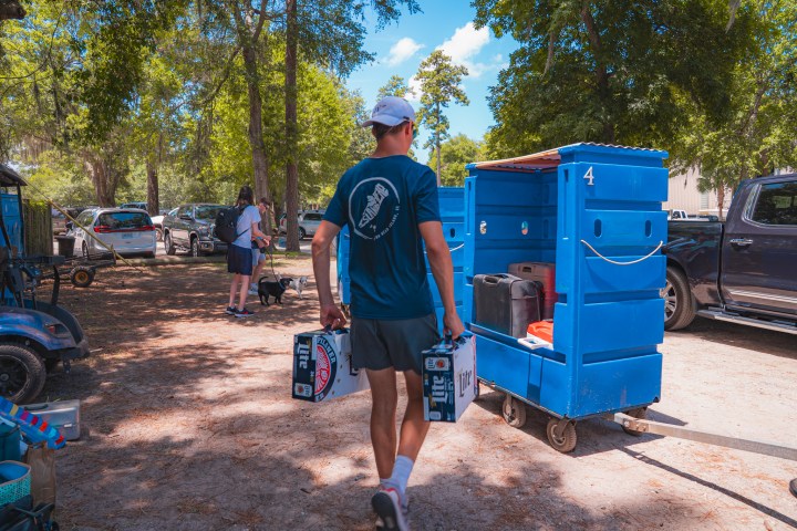 Person wearing cap carrying drinks by a blue cart in a park with trees and cars.