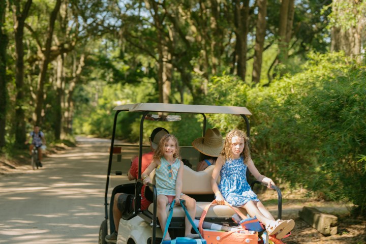 Children riding on the back of a golf cart with bags, on a tree-lined road.