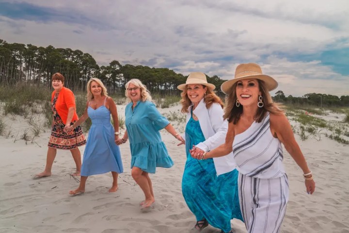 Five smiling women in summer dresses walk hand in hand on a sandy beach.