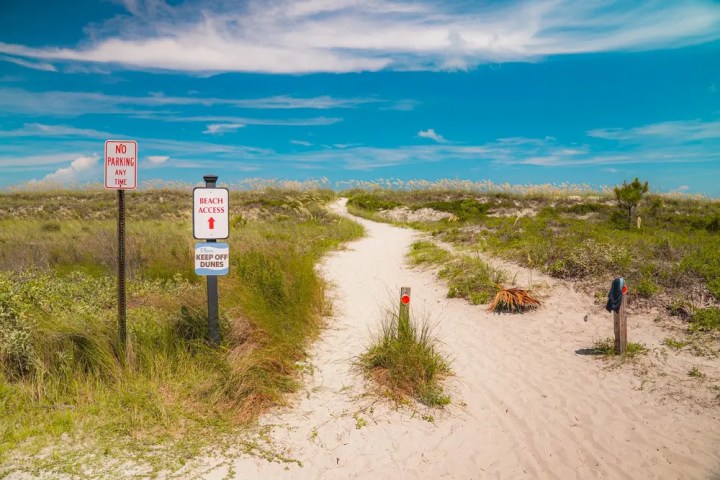 Sandy path leading to a beach with signage and grassy dunes under a blue sky.